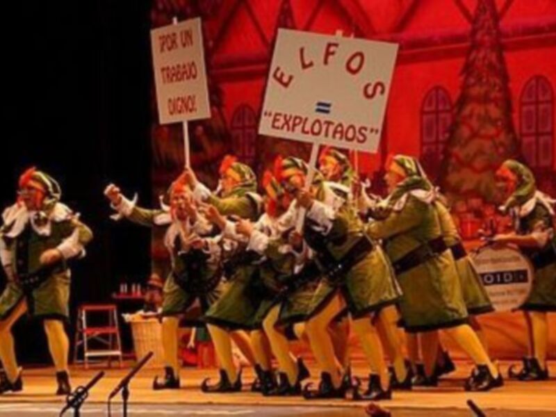 Un grupo de personajes vestidos de elfos en una escena teatral, con carteles que dicen 'ELFOS' y 'EXPLOTAOS'.