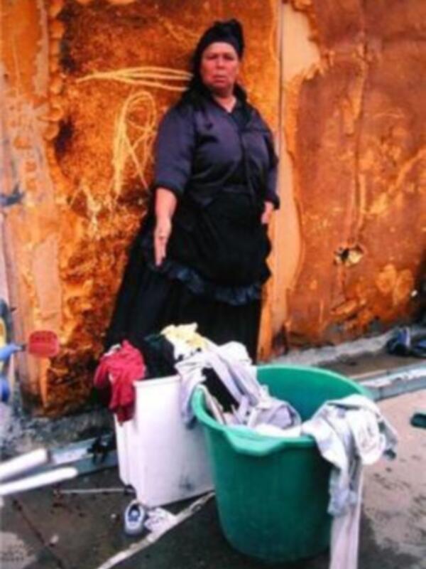 Una mujer vestida de negro con una gorra negra está frente a un muro oxidado. En el suelo, hay dos cubos de lavandería: uno blanco y otro verde con ropa sucia.