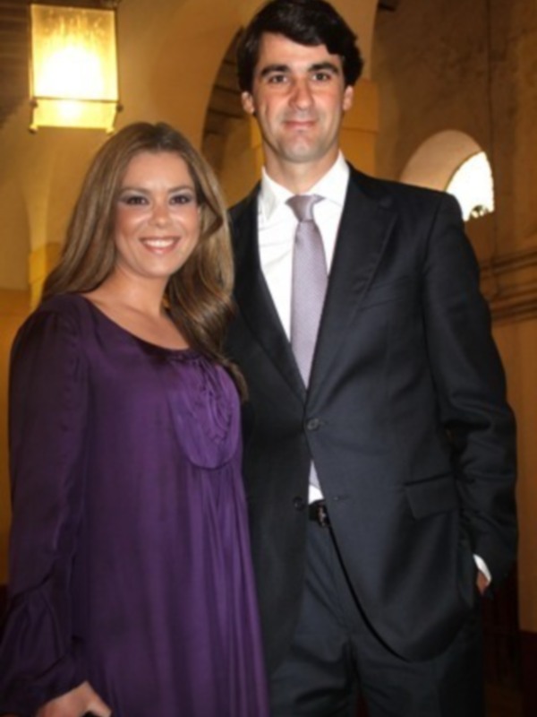 Una pareja elegante posa juntos en un ambiente formal, con una mujer en un vestido morado y un hombre en un traje oscuro.