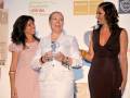 Tres mujeres sonrientes en una ceremonia de premiación, con un fondo de publicidad para 'custom drinks' y 'Barceló Premium'.