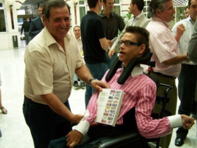 Una persona en silla de ruedas sostiene un folleto mientras un hombre le ayuda. Contexto: evento o conferencia, gente en el fondo.