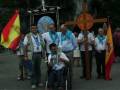 Una procesión religiosa con personas vestidas de blanco, algunos con banderas españolas y un hombre en silla de ruedas portando una cruz.