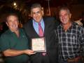 Tres hombres celebran una entrega de reconocimiento con un certificado en una noche al aire libre.
