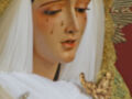 Una figura de la Virgen María con cabello blanco y una expresión triste, llorando. Está vestida con un manto blanco y lleva una corona dorada en la cabeza, con detalles florales. La escena parece ser de un altar o iglesia.