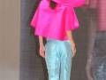 Una modelo camina por la pasarela, vestida con una chaqueta rosa brillante y pantalones azules claro. La imagen está enfocada en la parte trasera de la modelo, con un fondo que parece ser una pantalla digital.