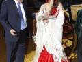 Una mujer vestida con un traje flamenco rojo y blanco posa junto a un hombre en traje formal. Detrás de ellos, se observa una carroza clásica con ruedas grandes y un fondo dorado.