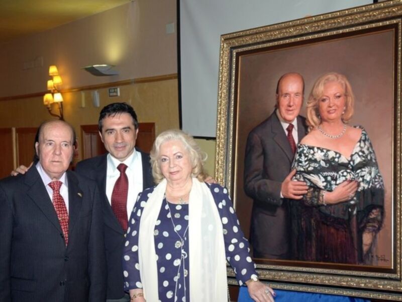 Dos personas posan junto a una pintura de un matrimonio enmarcado. La escena parece ser en un evento formal, posiblemente una recepción o conferencia.