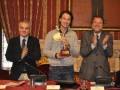 Tres hombres celebran en una sala de conferencias, uno sostiene un trofeo mientras los otros aplauden. La decoración y el ambiente sugieren una ceremonia formal o de reconocimiento.