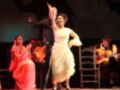 Una pareja baila flamenco en el escenario, mientras un músico toca guitarra y otro toca batería. La pareja está en el centro, con la bailarina levantando una mano y el bailarín apoyado en un pie. La música se puede escuchar a través de los instrumentos.