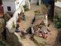 Un pequeño pueblo en miniatura con casas blancas y verdes, figuras humanas pequeñas y animales en un entorno de arena.