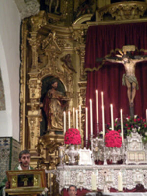 Altar con estatua de San Juan Evangelista, candelabros y flores rojas.