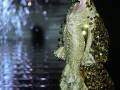 Model wearing a stunning gold dress with intricate designs, walking on a wet runway with fireworks in the background.