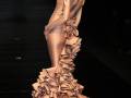 Una modelo presenta una vestimenta de gala con un vestido largo y elegante, adornado con flores en la falda. La pieza se complementa con un sombrero y joyas, destacando su estilo sofisticado.
