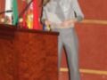 La Reina Letizia de España, con un vestido gris y pantalones, se encuentra en una conferencia o evento público. Detrás de ella hay banderas españolas y andaluzas, lo que sugiere una celebración o aniversario.