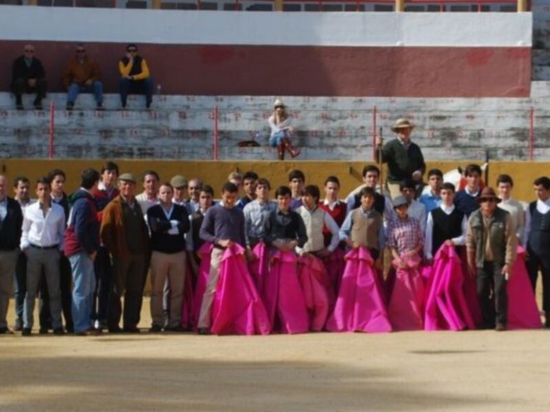 Grupo de jóvenes y adultos posando en un ruedo de toros con trajes de flamenca y trajes típicos, en un evento tradicional.