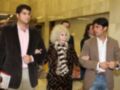 Dos hombres jóvenes caminan junto a una mujer mayor con un abrigo estampado. La escena parece ser en un entorno formal, posiblemente una sala de conferencias o evento.