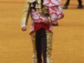 Un torero en traje tradicional de color dorado y rojo, con un sombrero de copa, camina en la plaza de toros. La arena es roja y el fondo muestra una estructura metálica.