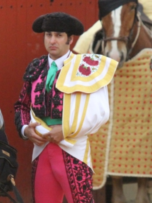 Un torero en traje de luces durante una corrida de toros, con un caballo y el fondo de una plaza de toros roja.