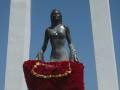 Una estatua de bronce en un pedestal blanco, con una corona en la parte superior. La estatua sostiene una cesta roja llena de flores y un collar dorado. El cielo azul claro se ve en el fondo.