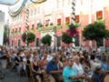Una multitud asiste a una celebración frente a un edificio histórico con ventanas y balcones decorados. La gente está sentada en sillas, disfrutando de la fiesta.