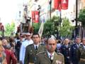 Desfile militar en una calle con banderas y personas en el fondo.}