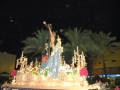 Imagen nocturna de una procesión religiosa con estatuas y flores en un carro. Se observan palmeras y edificios en el fondo, iluminación festiva y un cielo oscuro.