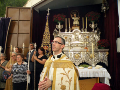 Un sacerdote en vestimenta religiosa, con una cruz y flores en el fondo.