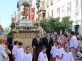 Una procesión religiosa con niños vestidos de blanco y rojo, acompañados por adultos vestidos de negro, caminan por una calle con edificios históricos en el fondo.