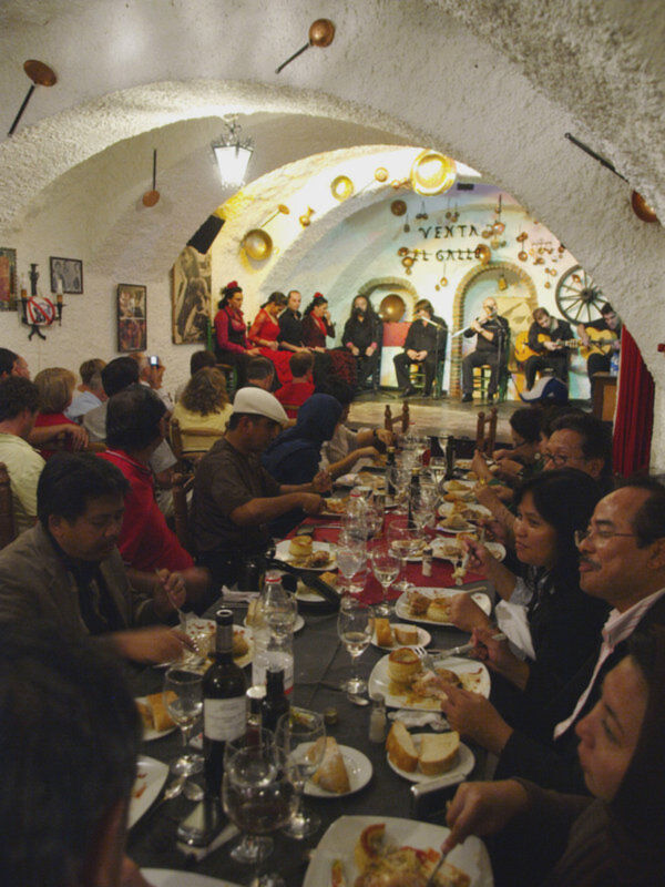 Una escena en un restaurante con una mesa larga llena de comidas y bebidas. Los clientes disfrutan de su comida mientras un grupo de músicos toca en el fondo, con una decoración festiva que incluye globos y un cartel que dice "VENTA". El ambiente es animado, con gente sentada en mesas y viendo a los músicos.