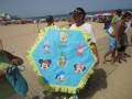 Un niño sostiene un sombrero de playa con dibujos animados famosos como Minnie Mouse, Mickey Mouse, Donald Duck y otros personajes de Disney. La playa está llena de gente disfrutando del sol y el mar en un día soleado.