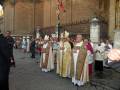Un grupo de sacerdotes vestidos con trajes ceremoniales caminan por una calle, frente a un edificio histórico. La imagen muestra detalles de la vestimenta religiosa y el ambiente urbano.