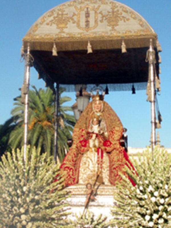 Una estatua de una Virgen María vestida en trajes dorados y rodeada por flores. Se encuentra en un lugar con palmeras y cielo azul claro, posiblemente durante una procesión religiosa.