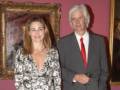 Una pareja posa frente a un retrato de pintura en una sala de exposiciones. La mujer lleva un vestido floral y el hombre, un traje gris con corbata roja. La pared de fondo está adornada con cuadros y el ambiente sugiere una celebración o evento cultural.