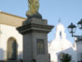 Estatua dorada de la Virgen en una columna de piedra, frente a una iglesia blanca con una cruz en la cúpula. Un grupo de personas se encuentra en el fondo, observando la escena.
