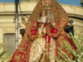 Imagen de una Virgen María vestida con trajes coloridos y joyas, posiblemente en un desfile religioso o procesión.