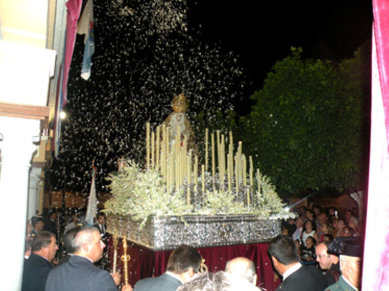 Una procesión nocturna con una imagen religiosa en un carro adornado con velas y flores, rodeada de personas observando la escena.