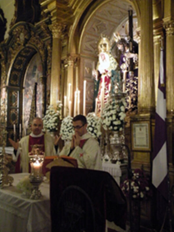 En una iglesia, dos sacerdotes vestidos con hábitos blancos y rojos se encuentran frente a una imagen de la Virgen María en un altar ornado. Alrededor, se pueden ver velas encendidas y flores blancas.