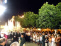 Fiesta religiosa en una plaza con una procesión nocturna, personas vestidas formalmente y árboles en primer plano.