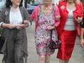 Tres mujeres caminan juntas, una con pantalones y chaqueta, otra con vestido floral y otro con falda roja. En el fondo, se ve un edificio blanco y una calle con coches.