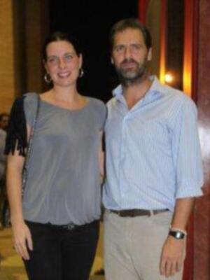 Dos personas posan juntas en un evento, uno con camisa azul y el otro con una blusa gris. Ambos llevan pantalones oscuros y la escena parece ser en un lugar con cortinas de color rojo.