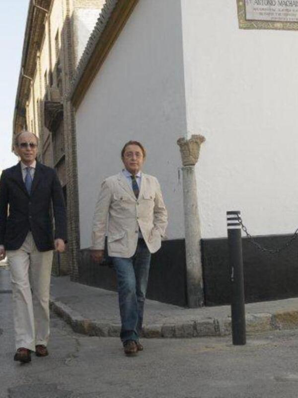 Dos hombres caminan por una calle adoquinada junto a un edificio blanco con una placa en la fachada.