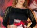 Una mujer con cabello largo y rojo, vestida de negro con detalles dorados, sostiene una revista con la portada de Jessica Bueno.