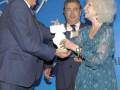 Una mujer con cabello blanco y una pieza de vestido azul está recibiendo un premio de manos de dos hombres. El fondo muestra una pantalla con el texto "VIII Premios" y "Matazo Tierras".