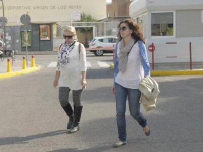 Dos mujeres caminan por una calle con un edificio y un coche en el fondo. Una lleva una bufanda y sombrero, mientras que la otra tiene un bolso beige.
