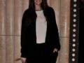 Una mujer con cabello largo y negro, vistiendo un traje negro y una camisa blanca. Posa con una mano en el bolsillo de su pantalón negro, sosteniendo un clutch con estampado animal. Fondo con cortinas y una puerta de cristal.