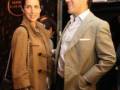 Una pareja en un evento formal, el hombre con una chaqueta beige y camisa blanca, la mujer con un abrigo marrón.
