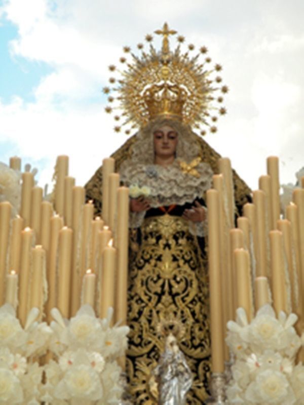 A una figura religiosa vestida con trajes dorados y negros, rodeada de velas blancas y flores. La figura lleva una corona dorada con estrellas y un manto bordado.