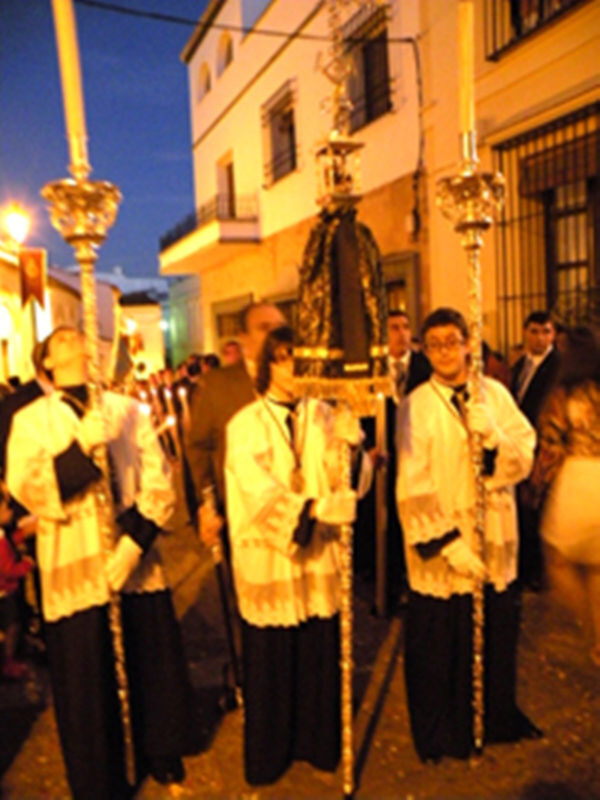 Una procesión religiosa en la noche, con personajes vestidos de blanco y negro, portando antorchas doradas.