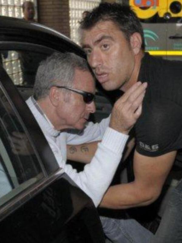 Un hombre en una camiseta negra y otro en una chaqueta blanca se abrazan mientras se sientan en un automóvil.