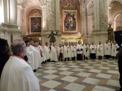 Imagen de un grupo de sacerdotes vestidos con hábitos blancos y rojos, posiblemente durante una ceremonia religiosa en un edificio histórico con arcos y vitrales.